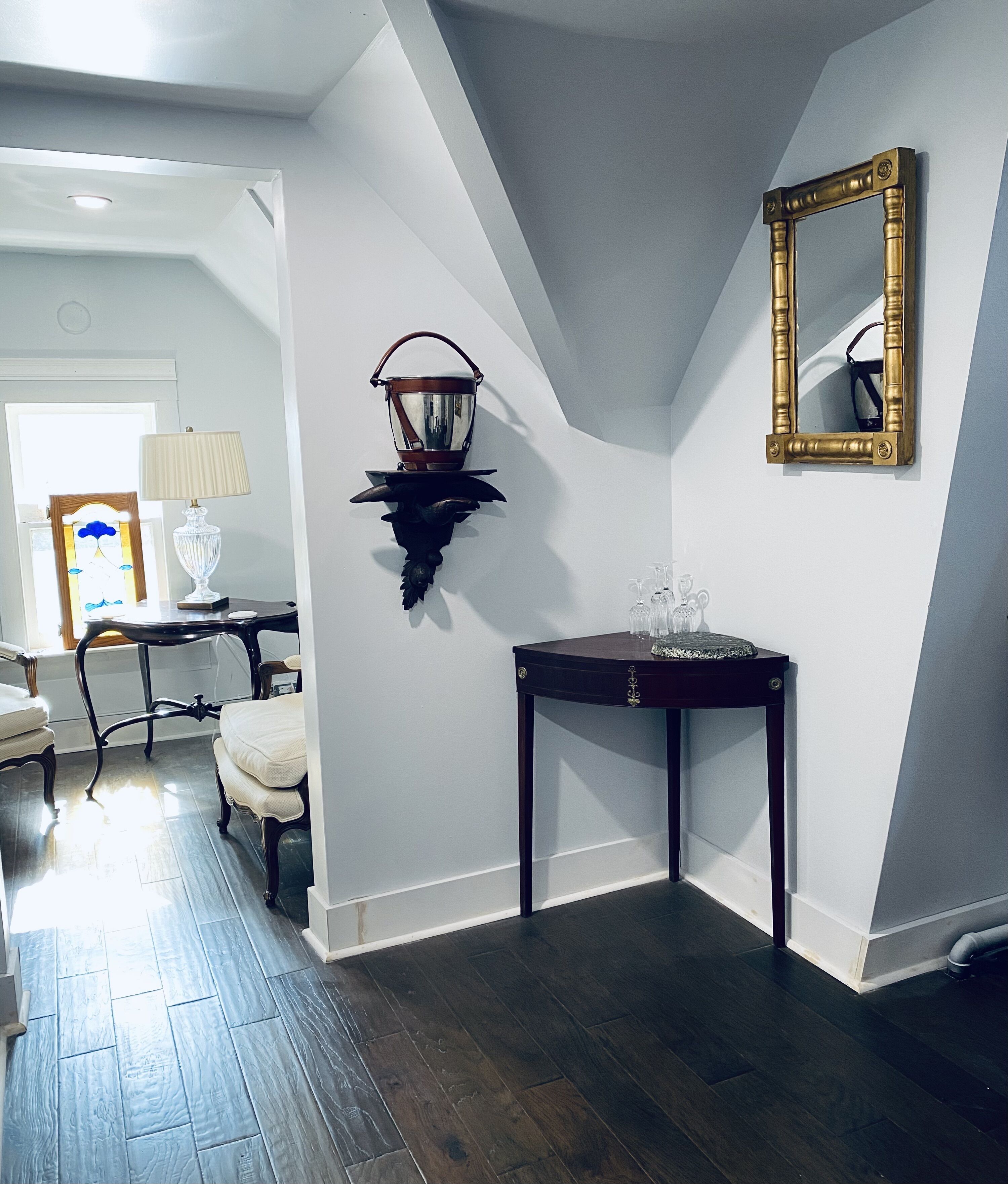Una habitación con suelo de madera oscura, pared blanca, consola de madera oscura con superficie de vidrio, lámpara de pared y un espejo.