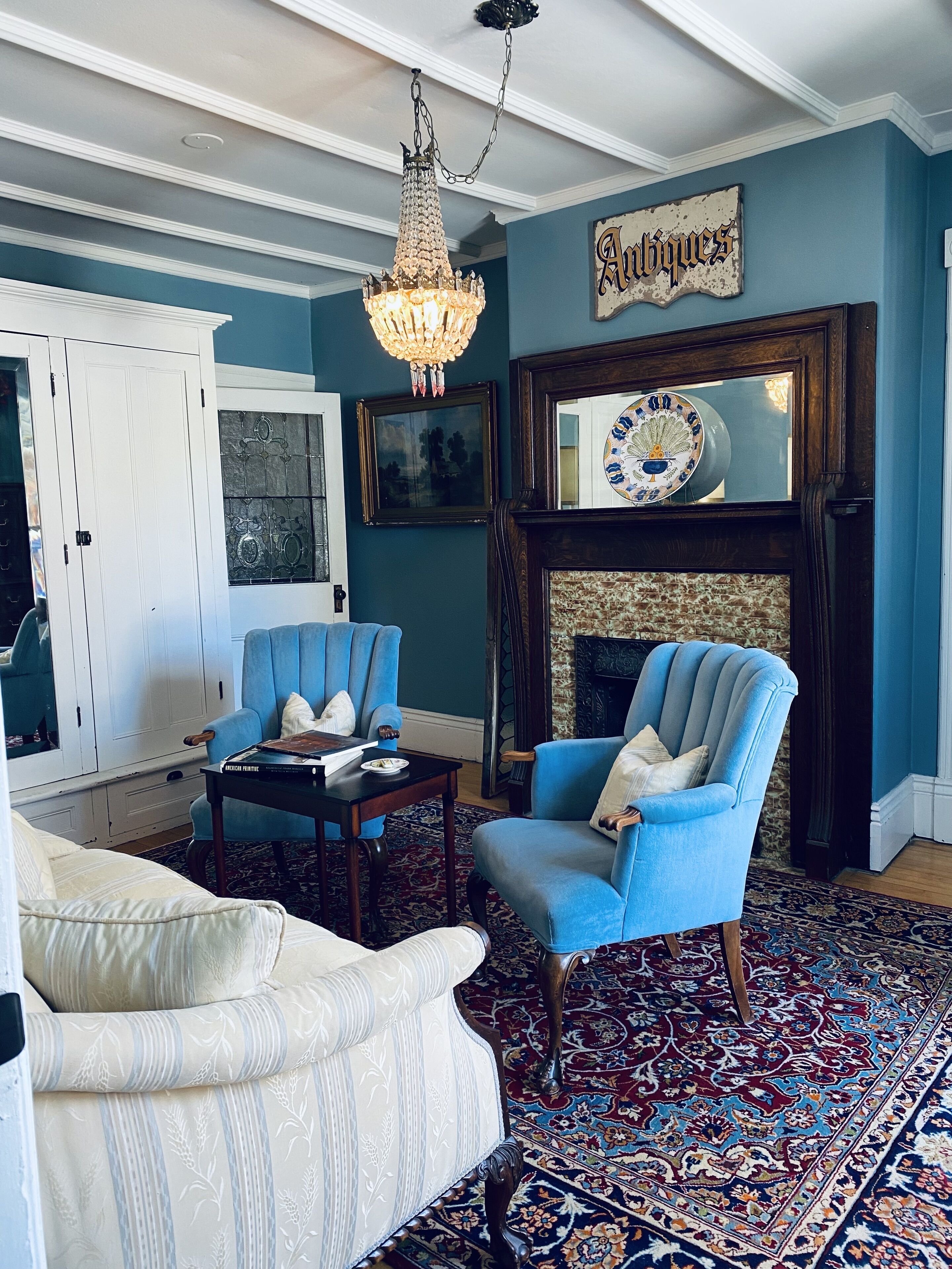 A living room with a blue armchair, a white armchair, a wooden coffee table, a chandelier, a fireplace, and a wall with a sign reading "Antiques".