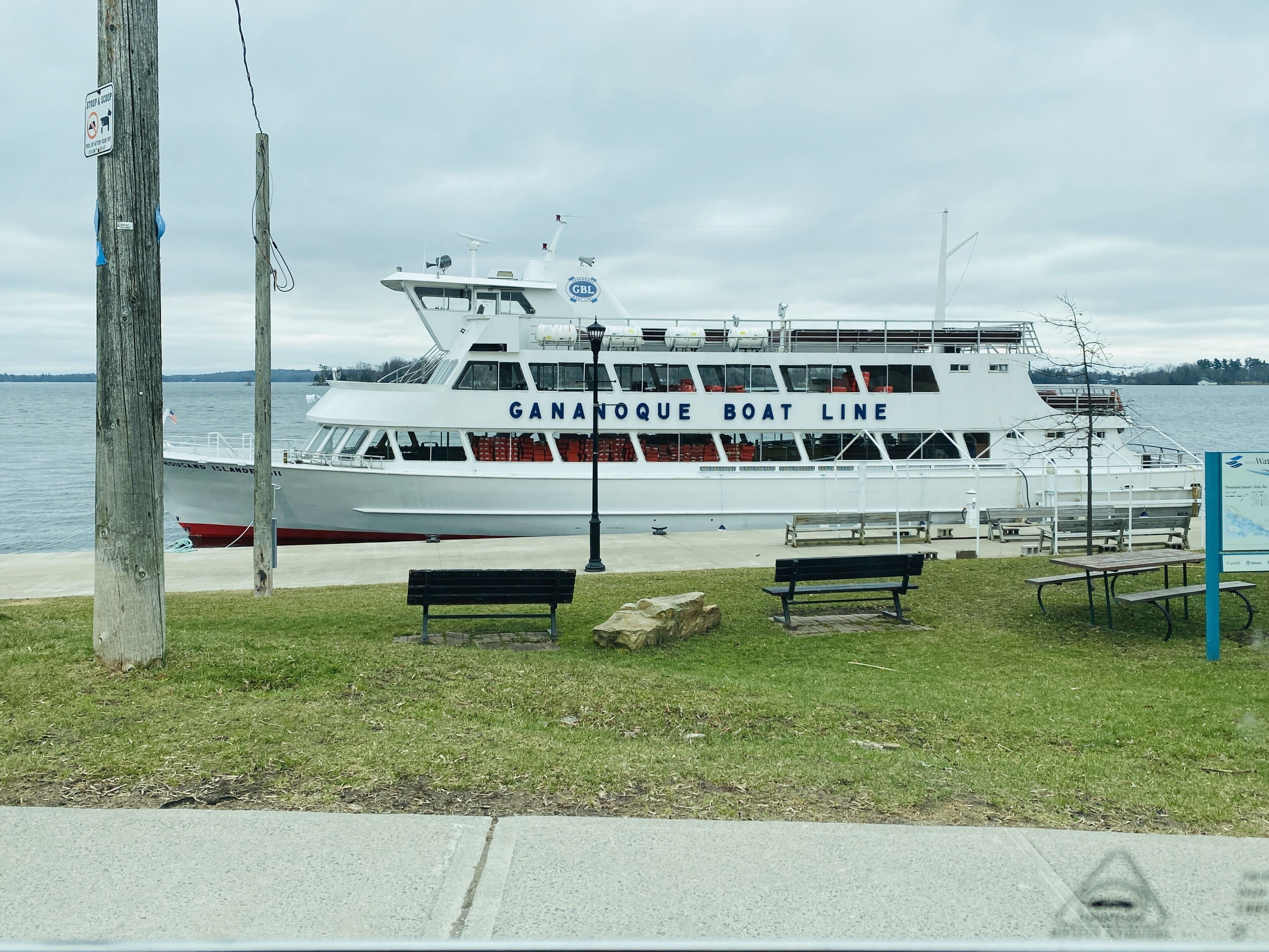 A white boat with "GANANQUE BOAT LINE" written on the side, docked at a pier with benches and picnic tables.