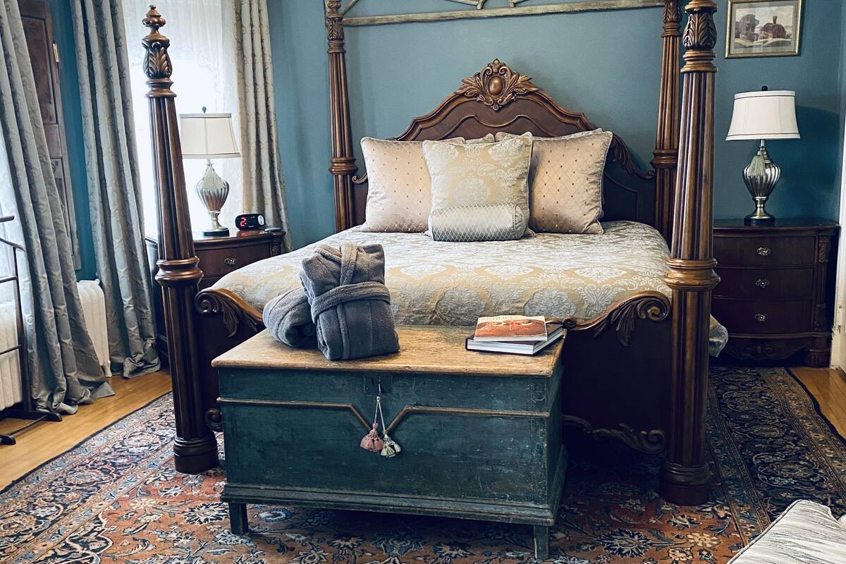 A bedroom with a four-poster bed, a nightstand with a lamp, a bedside table with a lamp, and a large window with curtains.