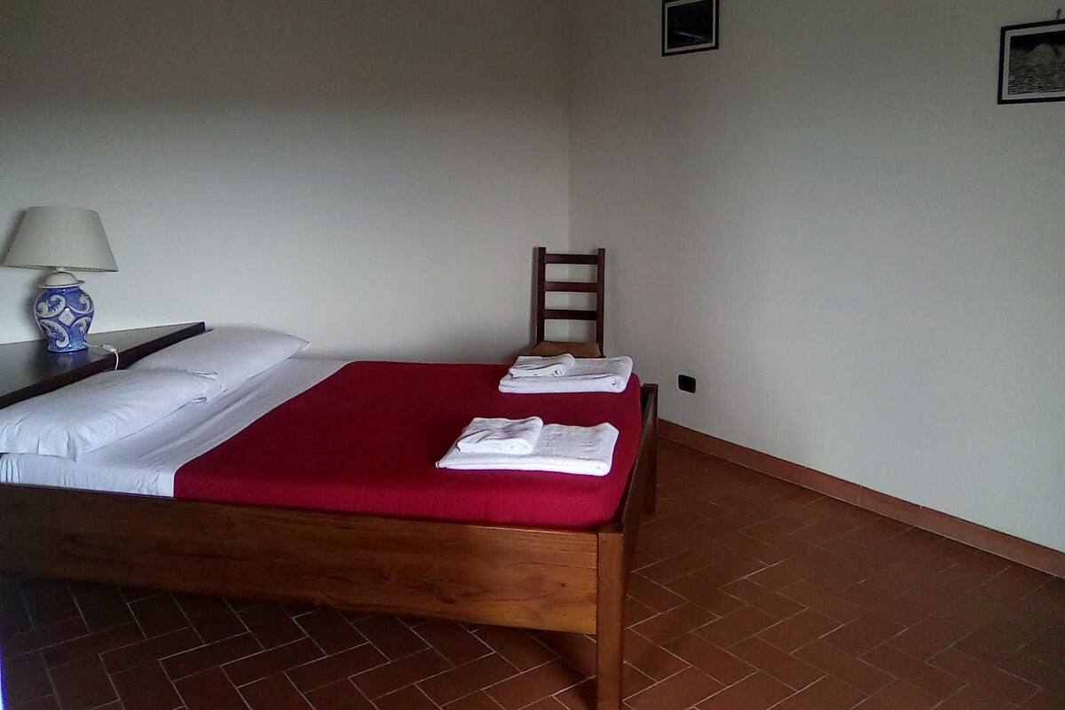 A single bed with a red bedspread, a wooden nightstand with a lamp, and a chair against the wall.