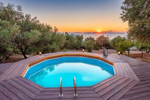 A swimming pool with a wooden deck, surrounded by trees, and a sunset view.
