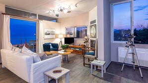 A cozy living room with a sofa, armchair, and a small table. There is a large window with a view of the outside. - Lagonisi Villa Afroditi (Saronikos)
