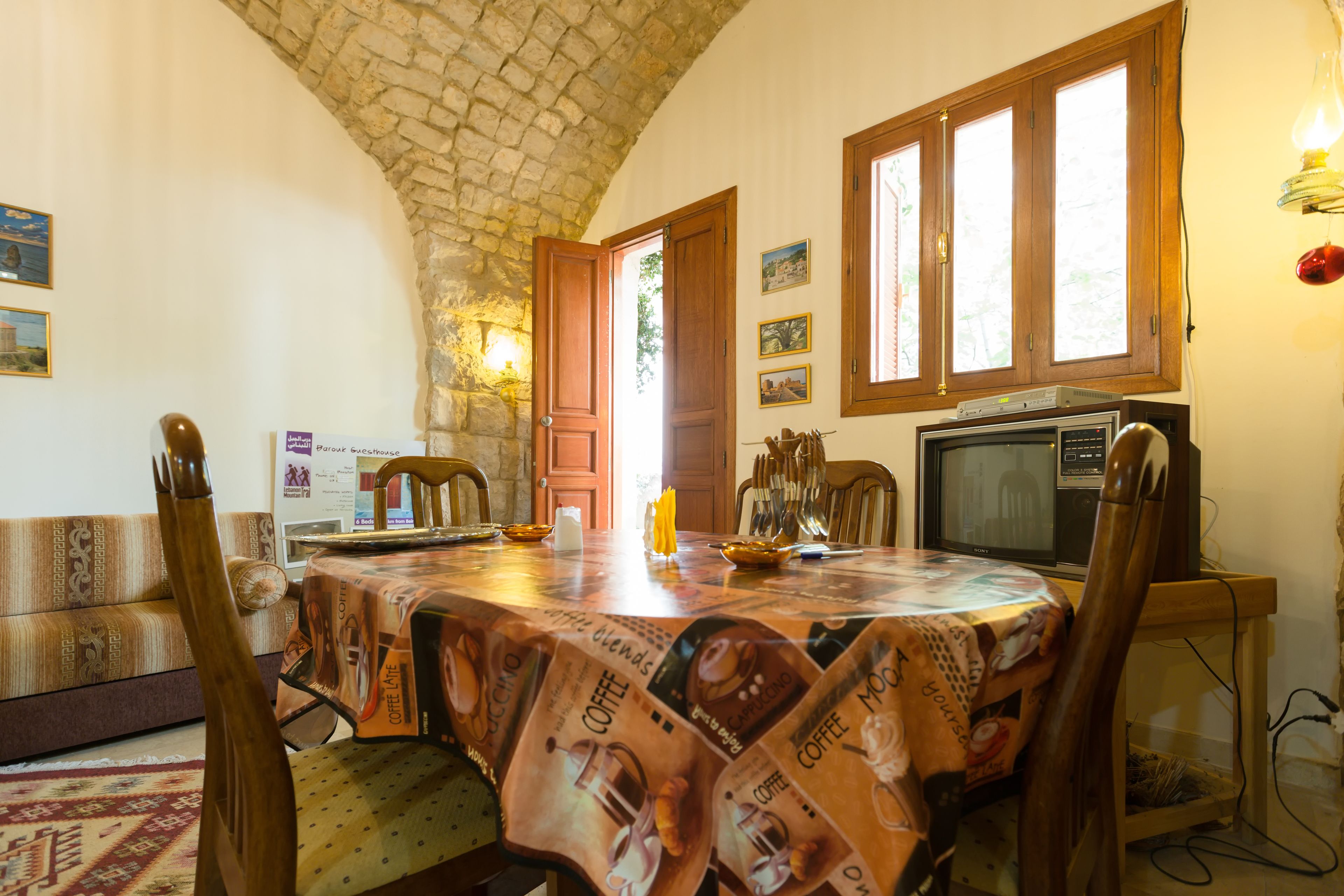 A dining area with a table covered in a coffee-themed tablecloth, wooden chairs, a television, and stone walls.