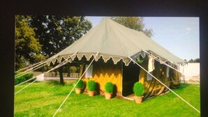 A large tent with a peaked roof and a scalloped edge, surrounded by a grassy area with potted plants. - Kumbha Kamps (Prayagraj)