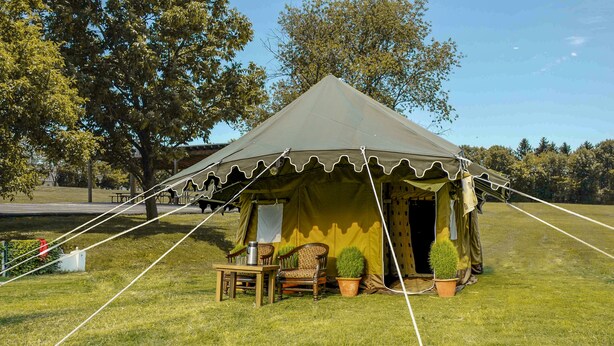 A tent with a table and chairs, surrounded by grass and trees. - Kumbha Kamps (Prayagraj)