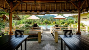 An outdoor seating area with wooden tables and chairs, umbrellas, and a view of a mountain and greenery. - Villa Manuk (Sawan)