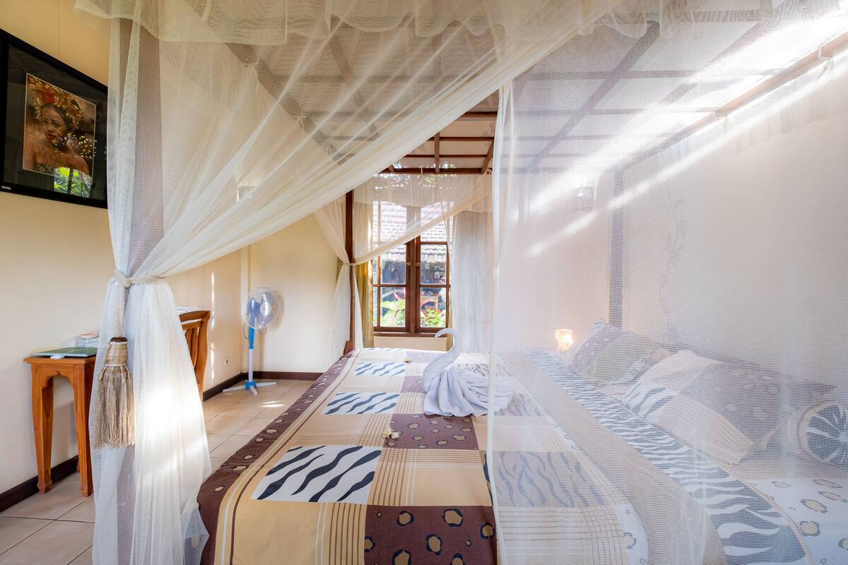 A room with a bed covered by a canopy, a wooden table, a standing fan, and a window with a view of greenery.