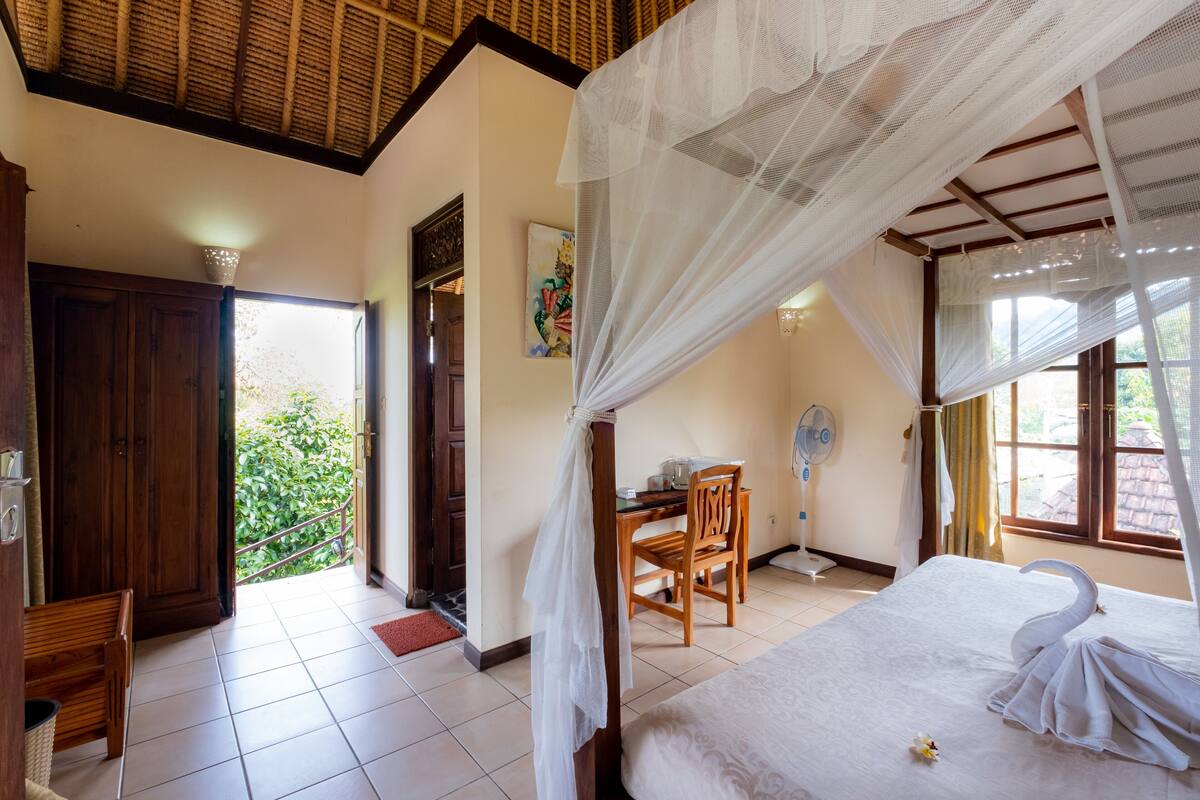 A room with a bed, a desk, a chair, a fan, and a view of greenery outside.