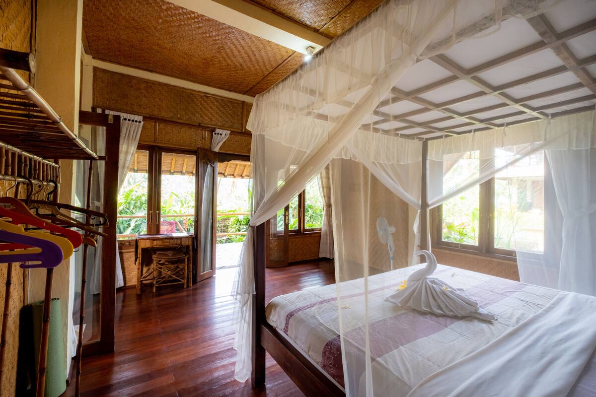 A room with a bed under a canopy, wooden flooring, a table with chairs, and a view of greenery outside.