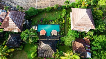 Vista aérea de un complejo turístico con una piscina central, rodeado de exuberante vegetación y varias edificaciones tipo bungalow con techos inclinados.