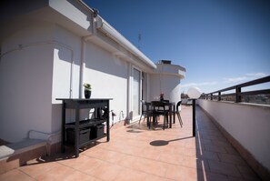 A rooftop terrace with a table and chairs, a small shelf with a potted plant, and a view of the city skyline.