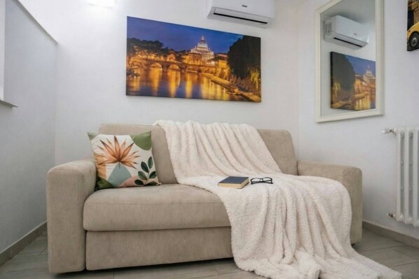 A beige sofa with a throw blanket and a pillow with a leaf pattern. There are two air conditioning units mounted on the wall.