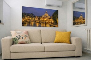 A neutral-toned sofa with two cushions, a wall-mounted air conditioner, and two framed pictures of a bridge and a building.