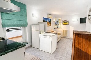 A compact kitchen with a dishwasher, a refrigerator, and a small island. There is a dining area with a sofa and a table. The room has a tiled floor and white walls.