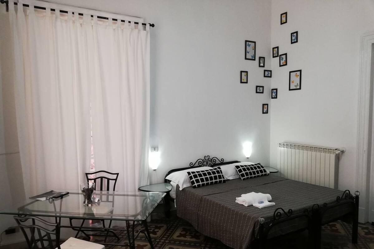 A bedroom with a bed, a glass table, chairs, a radiator, and wall-mounted pictures.