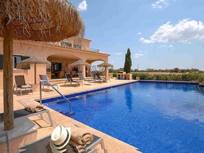 Área de piscina com espreguiçadeiras, um guarda-sol de palha e vista para o campo.