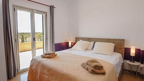 Quarto de hotel com uma cama grande, duas luminárias de cabeceira, uma mesinha e uma varanda com vista.