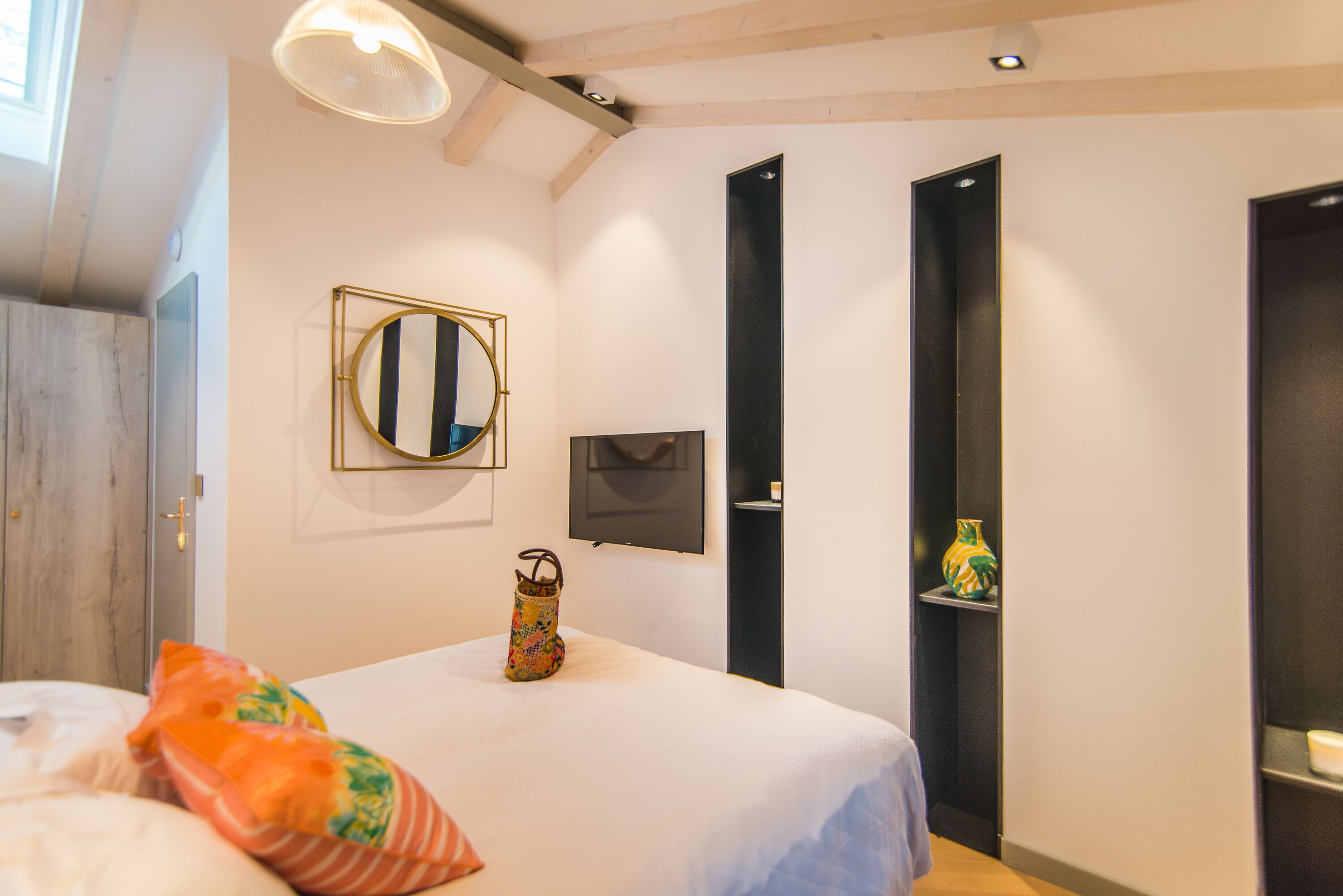 A bedroom with a bed, pillows, a mirror, a vase, and a television on the wall.