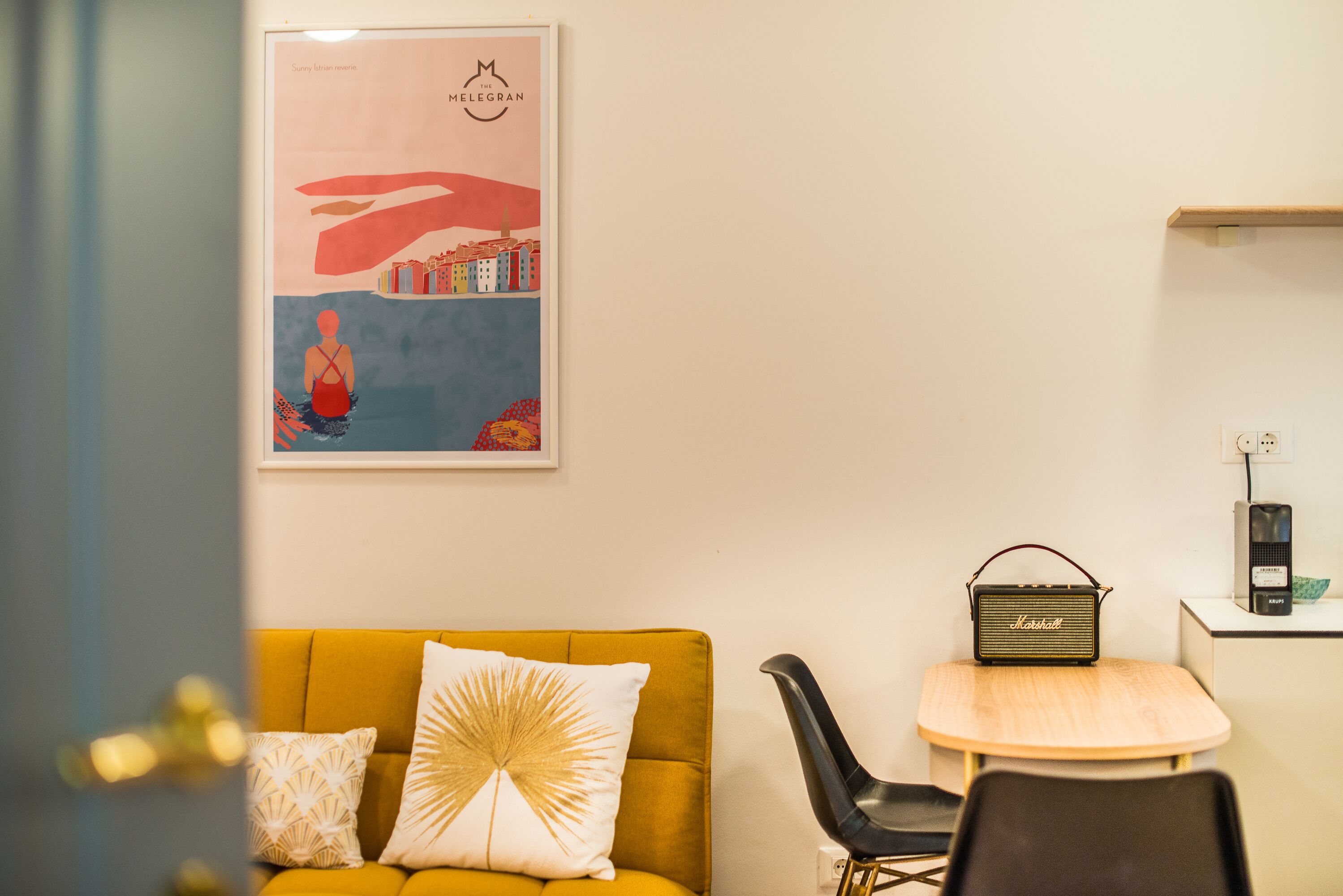 A room with a mustard yellow sofa, a white and gold patterned pillow, a black chair, a small round table, and a framed poster on the wall.