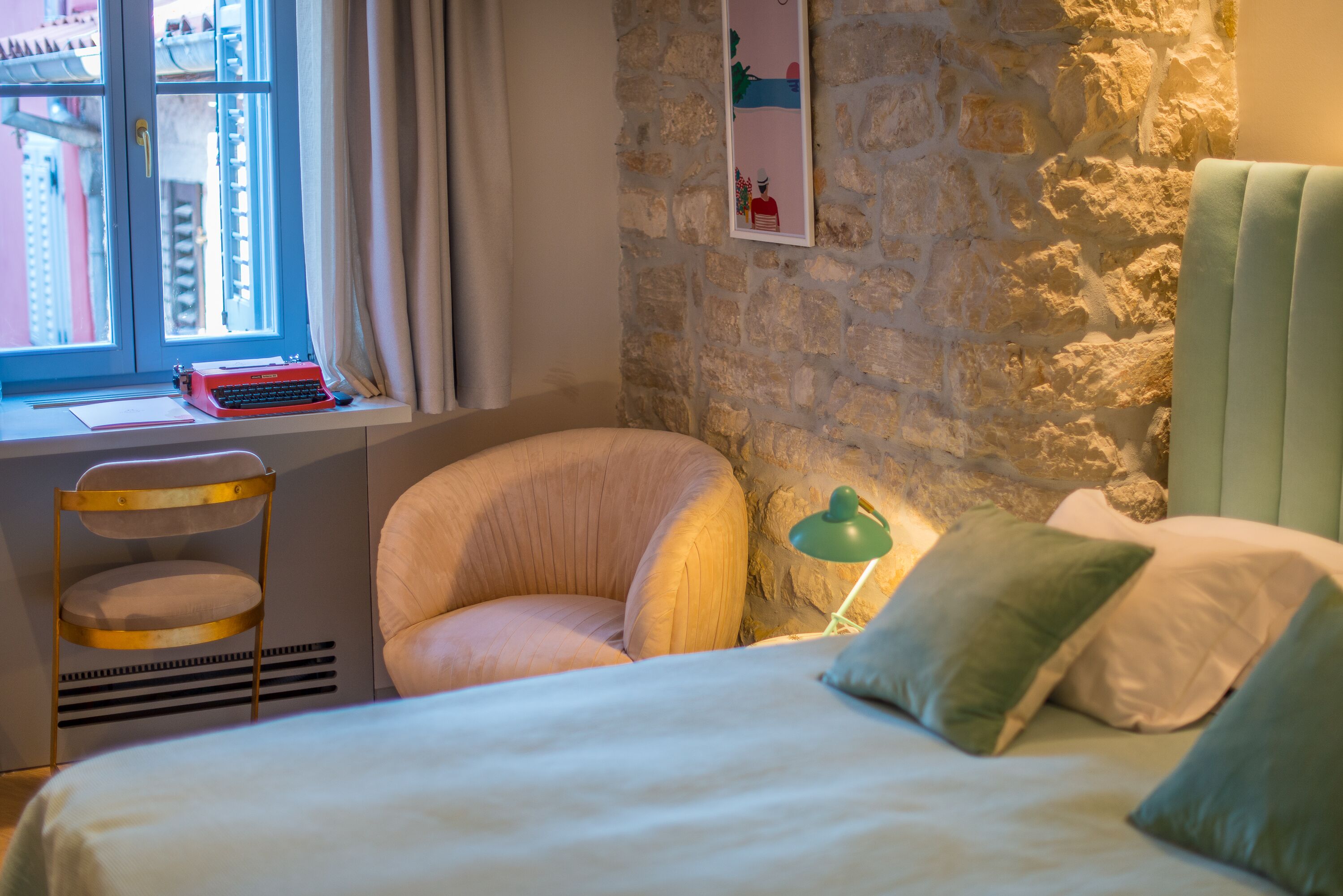 A bedroom with a stone wall, a window with curtains, a bed with pillows, a chair, a desk with a typewriter, and a lamp.