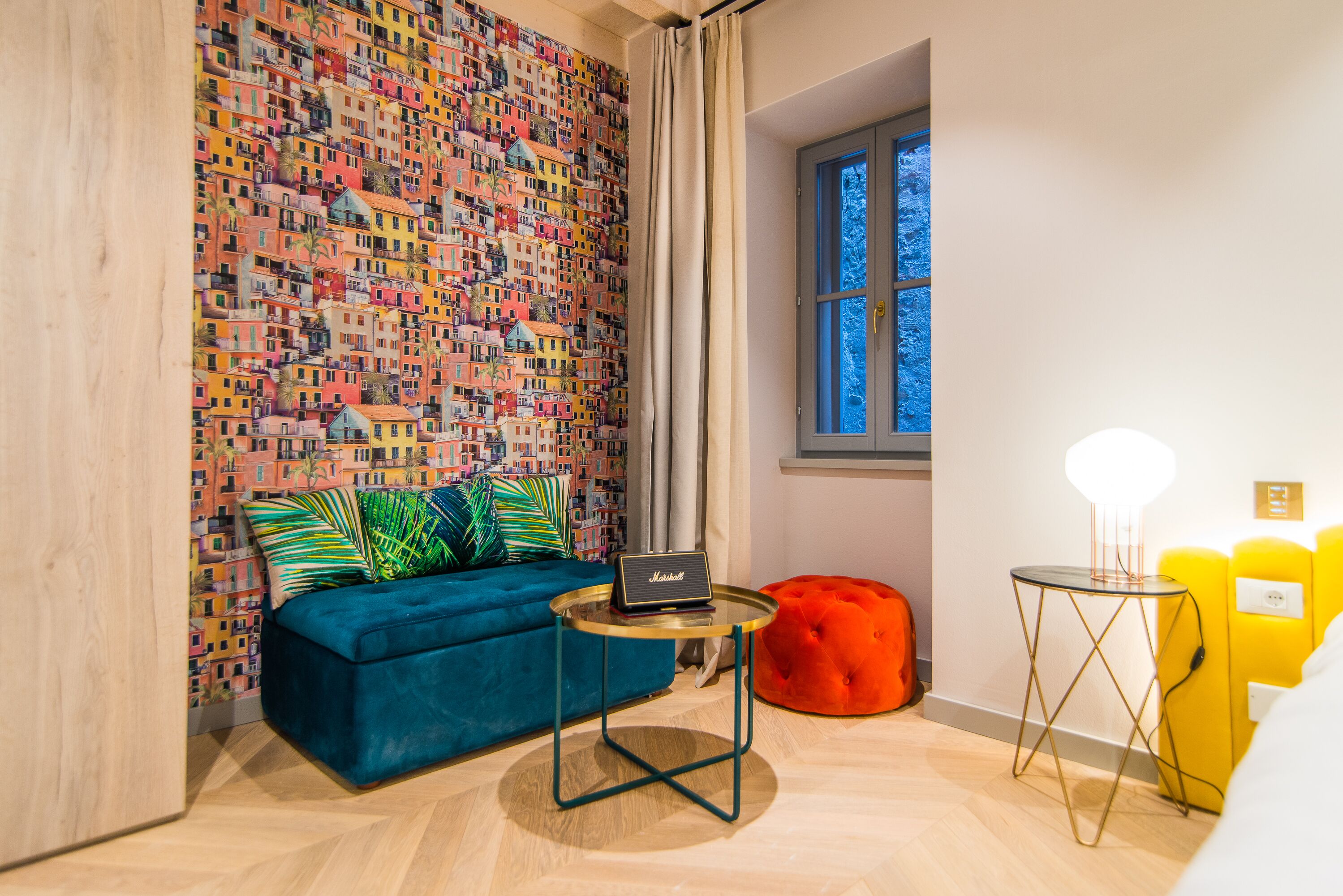 Ein Zimmer mit einer farbenfrohen Wandmalerei, einem blauen Sofa, einem kleinen runden Tisch mit einem Laptop, einem roten Hocker und einem Fenster mit Blick auf Bäume.