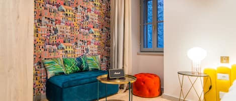 Une pièce décorée d’une fresque colorée, avec un canapé bleu, une petite table ronde sur laquelle se trouve un ordinateur portable, un pouf rouge et une fenêtre donnant sur des arbres.