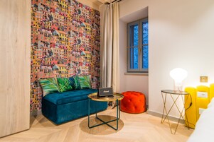 A room with a colorful mural, a blue sofa, a small round table with a laptop, a red ottoman, and a window with a view of trees.