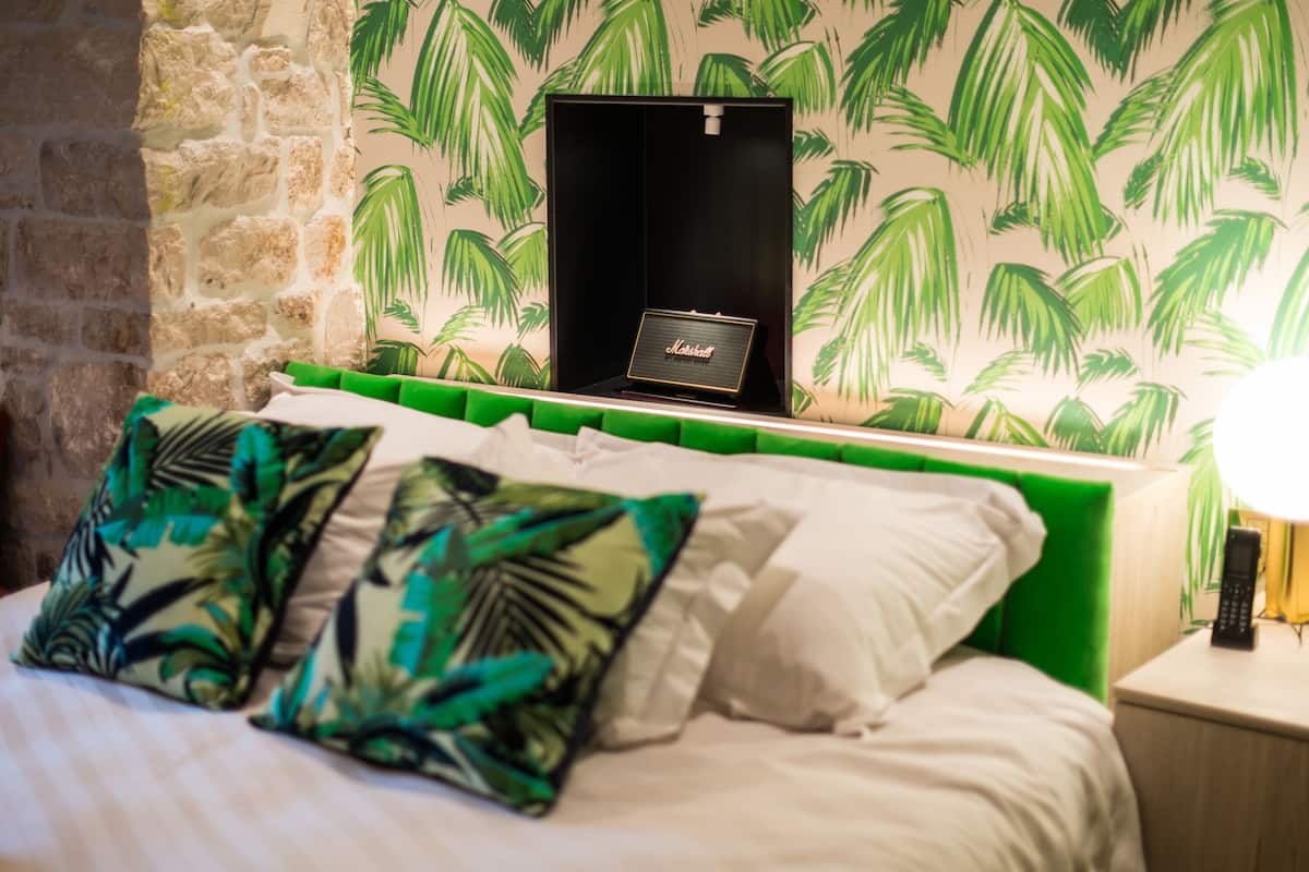 A bed with green headboard, white bedding, and tropical leaf-patterned pillows. A black box with a nameplate is on the headboard.
