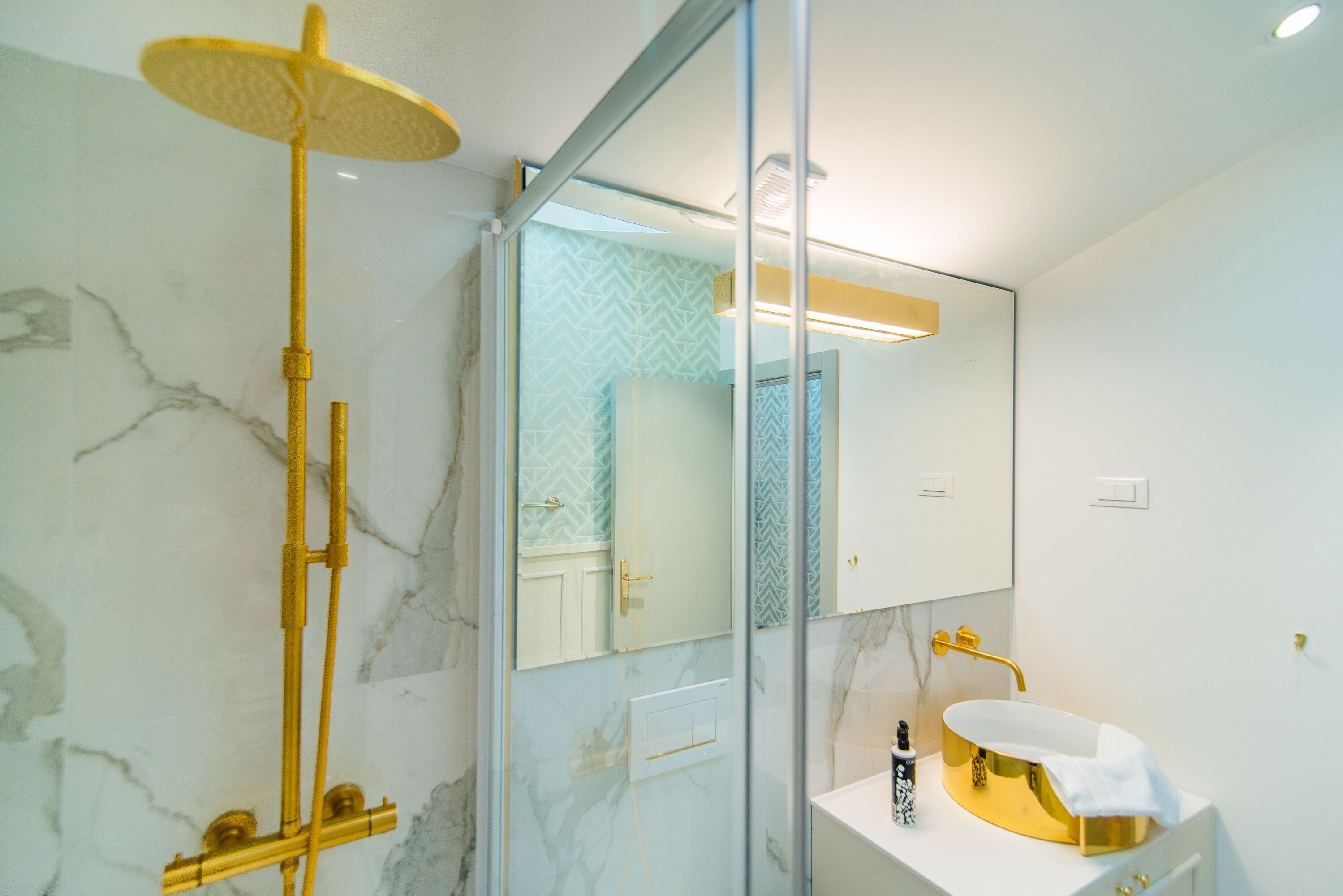 Un baño moderno con ducha de estilo dorado, un lavabo blanco con plato dorado y un espejo con un salpicadero geométrico.