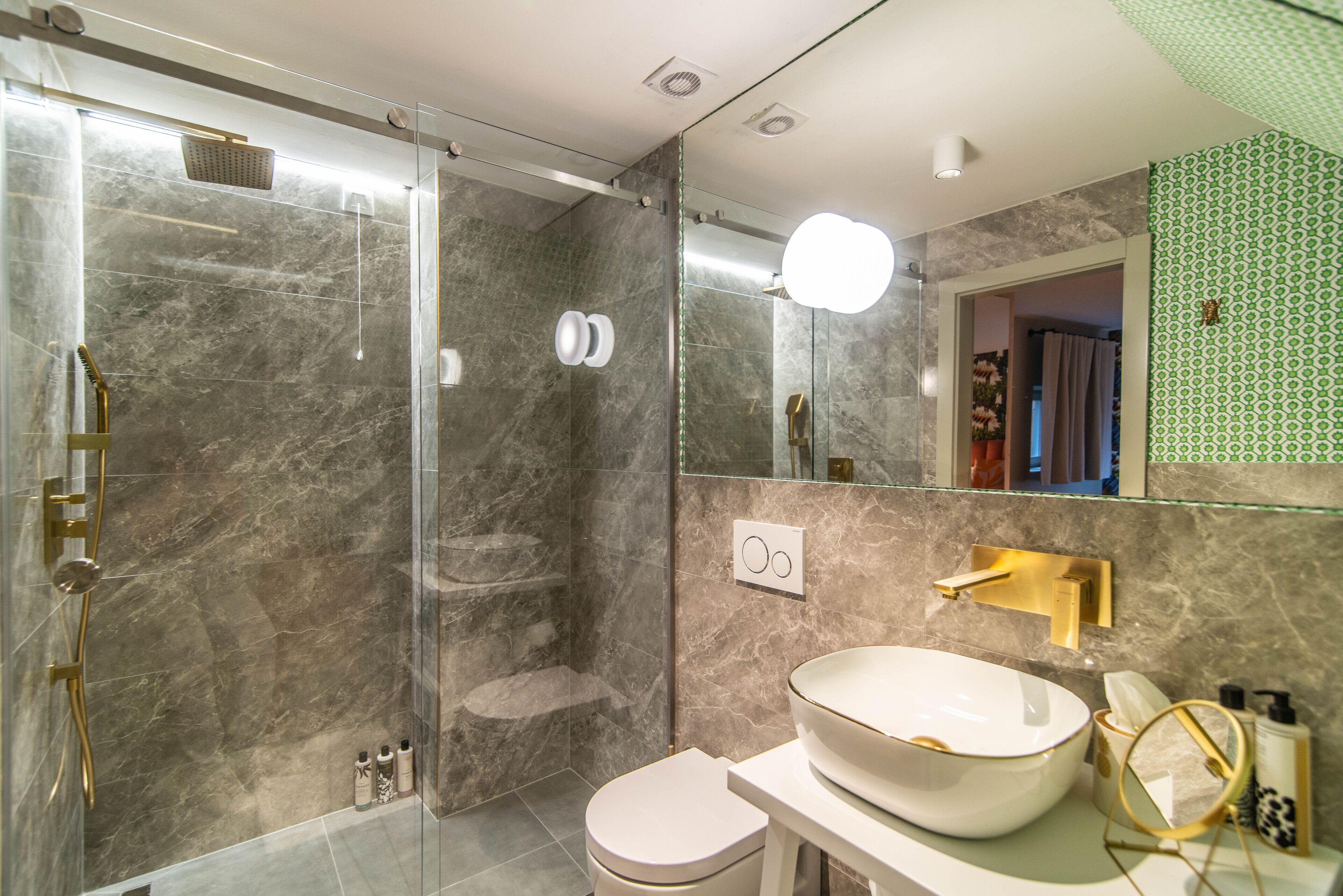 Een moderne badkamer met een douchecabine van glas, een witte wastafel met gouden accenten en een spiegel met een groene, gepatteerde lijst.
