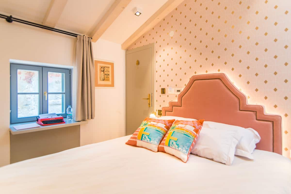 A neatly made bed with colorful pillows, a window with curtains, and a desk with a typewriter and a bottle.