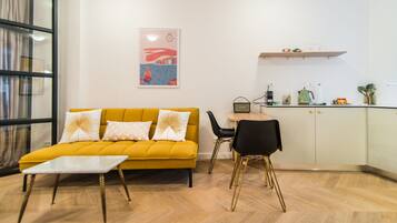 A modern living room with a yellow sofa, a small table, and a kitchen area with a dining table and chairs.