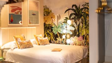 Una cama bien arreglada con ropa blanca y varias almohadas, un mural en la pared con plantas tropicales y paisaje urbano, y una lámpara colgante arriba.