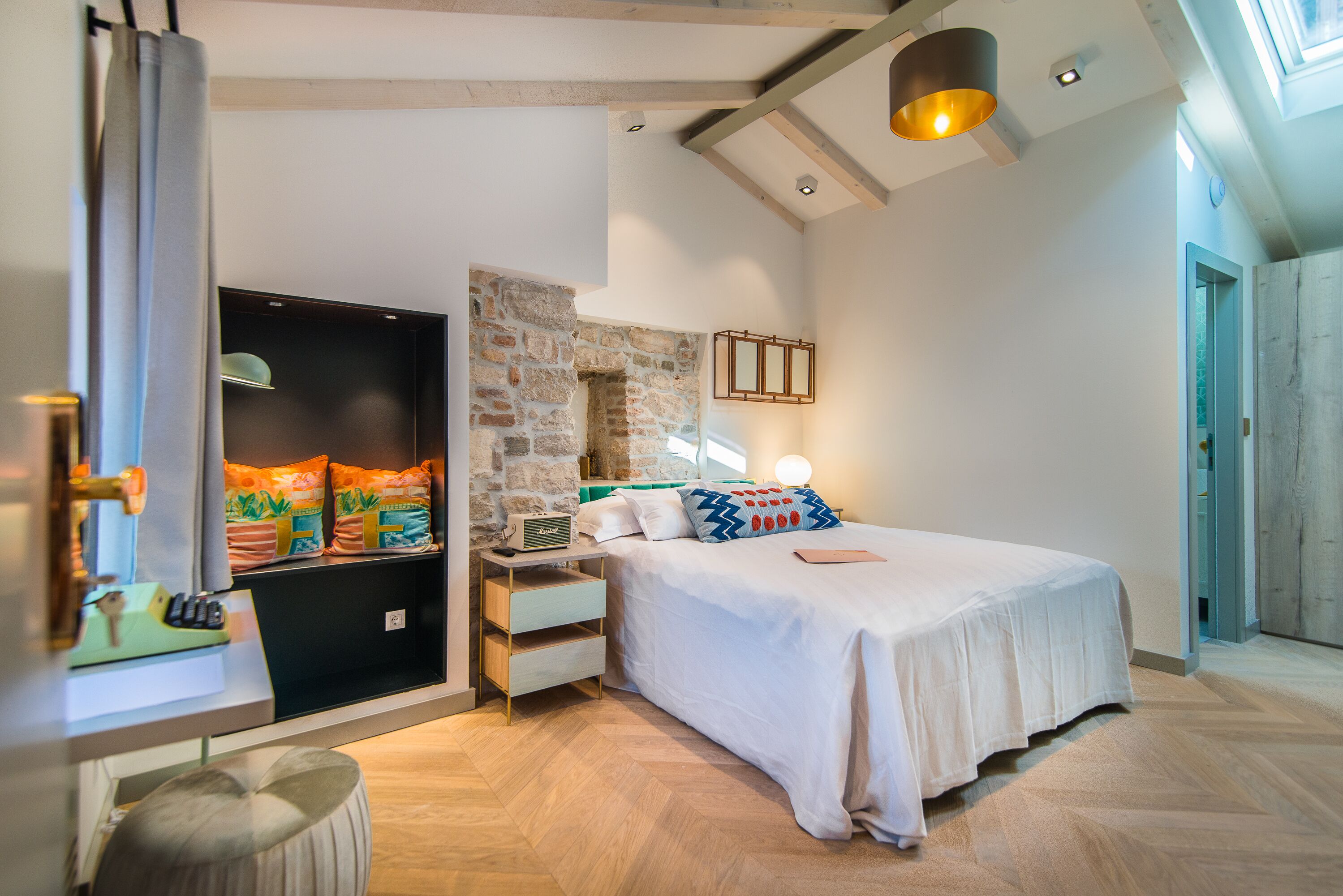 A bedroom with a bed, a nightstand, a lamp, a stone wall, and a wooden floor.