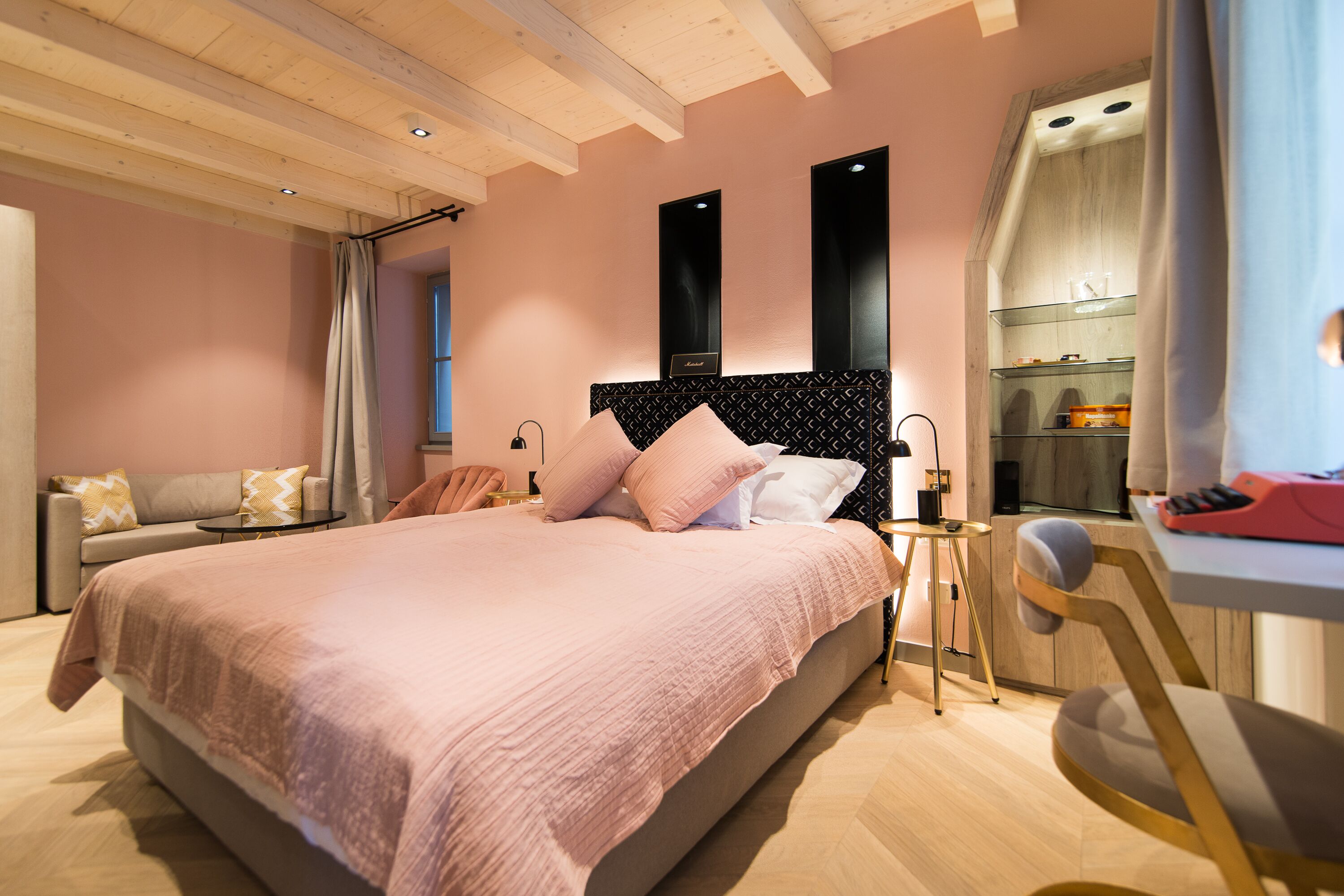 A modern bedroom with a large bed, bedside tables, a desk, and a kitchenette.