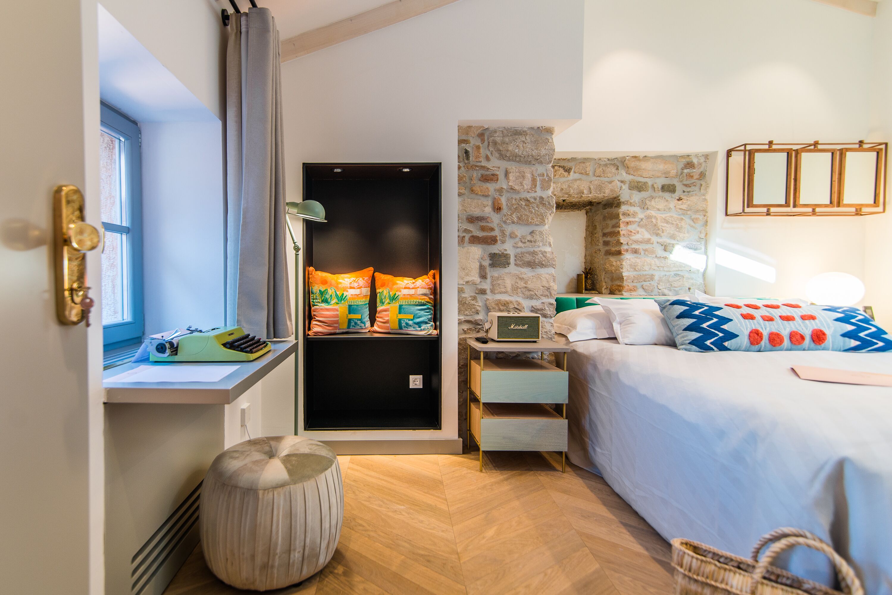 Un dormitorio con cama, mesita de noche, lámpara, ventana, puerta y pared de piedra.