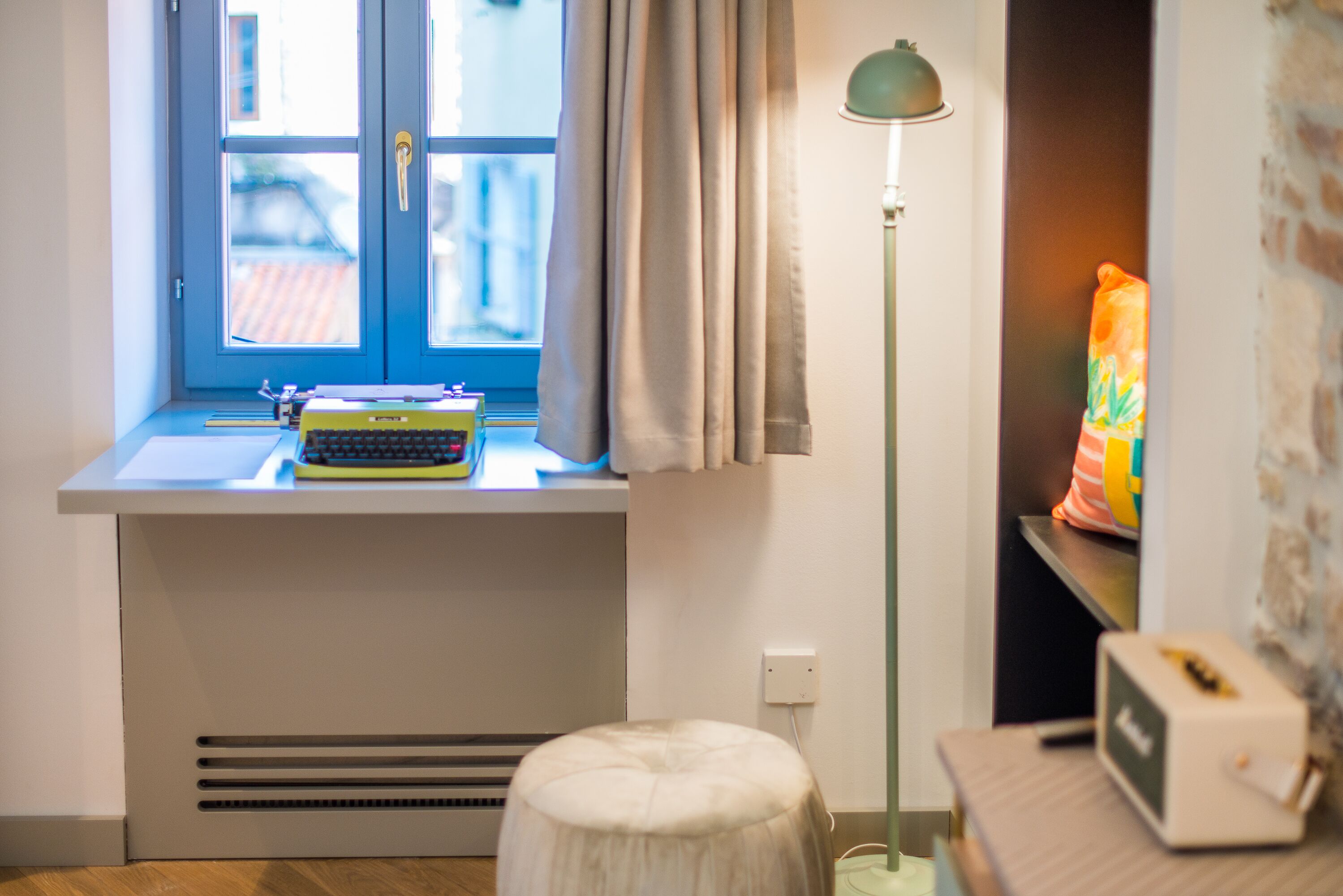 A room with a window, a desk with a typewriter, a lamp, and a small stool.