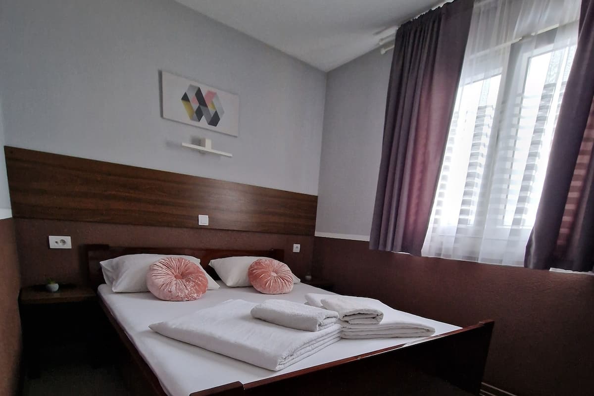 Habitación de hotel con una cama grande, cabecera de madera, mesita de noche pequeña y una ventana con cortinas.