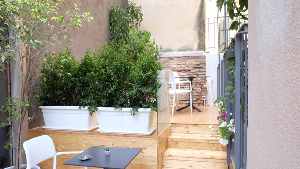 Una terraza de madera con sillas y una mesa blancos, rodeada de plantas en macetas y un muro de piedra.