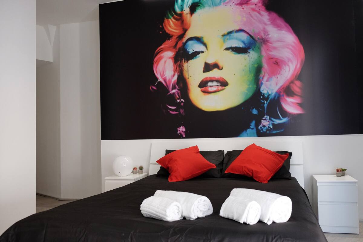 A modern hotel room with a large poster of a colorful artwork on the wall, a black bed with red and black pillows, and neatly folded white towels on the bed.