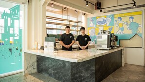 A modern kitchen area with two individuals standing behind a marble counter, a large window with a cityscape view, and a wall with colorful illustrations. - Meant to Be (Chiayi City)