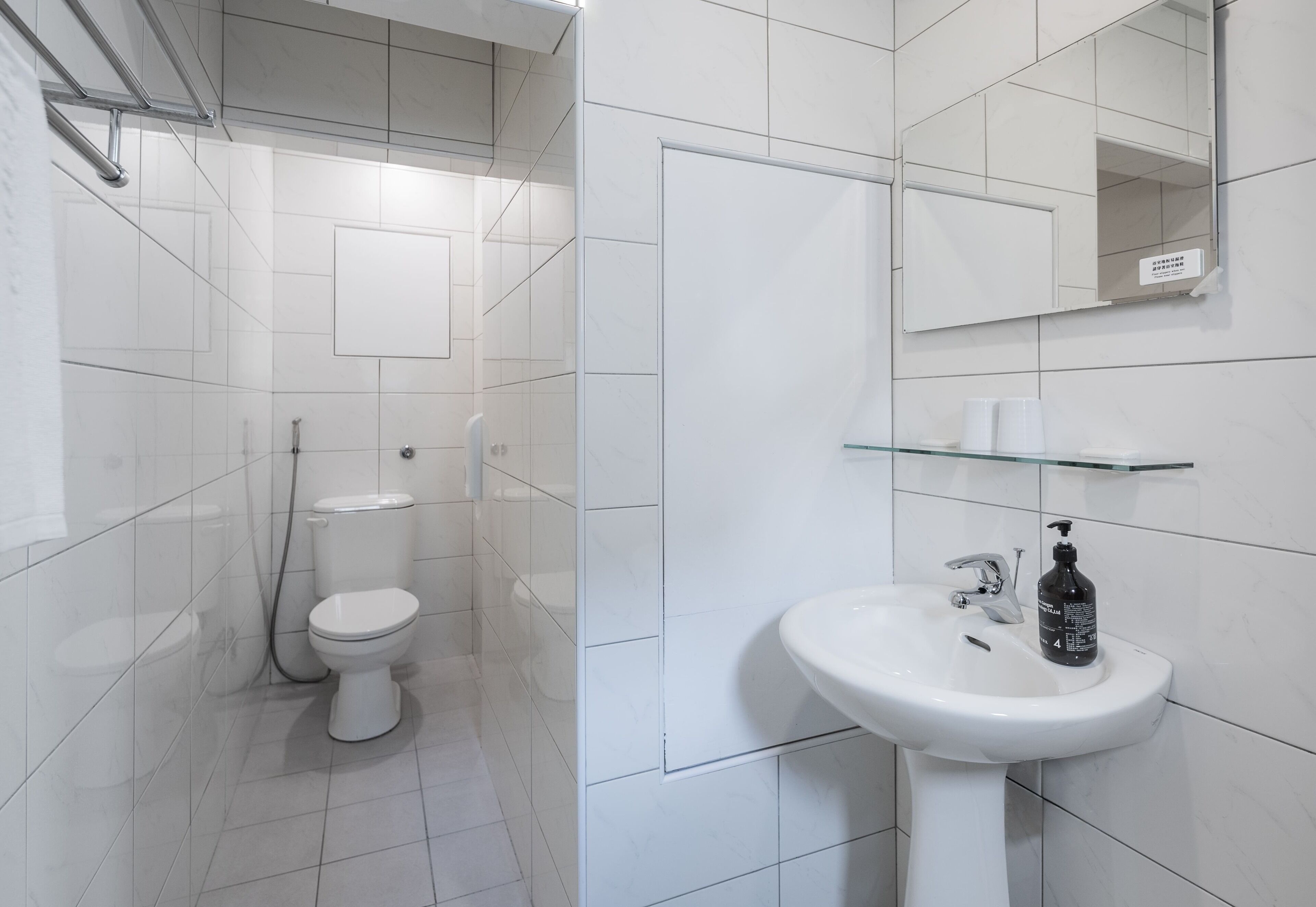 Un baño moderno con azulejos blancos, un lavamanos blanco, una repisa de vidrio y un inodoro.