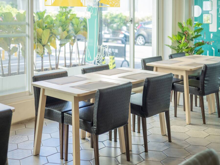 A restaurant interior with hexagonal tiled flooring, wooden tables and chairs, and large windows offering a view of the street outside.