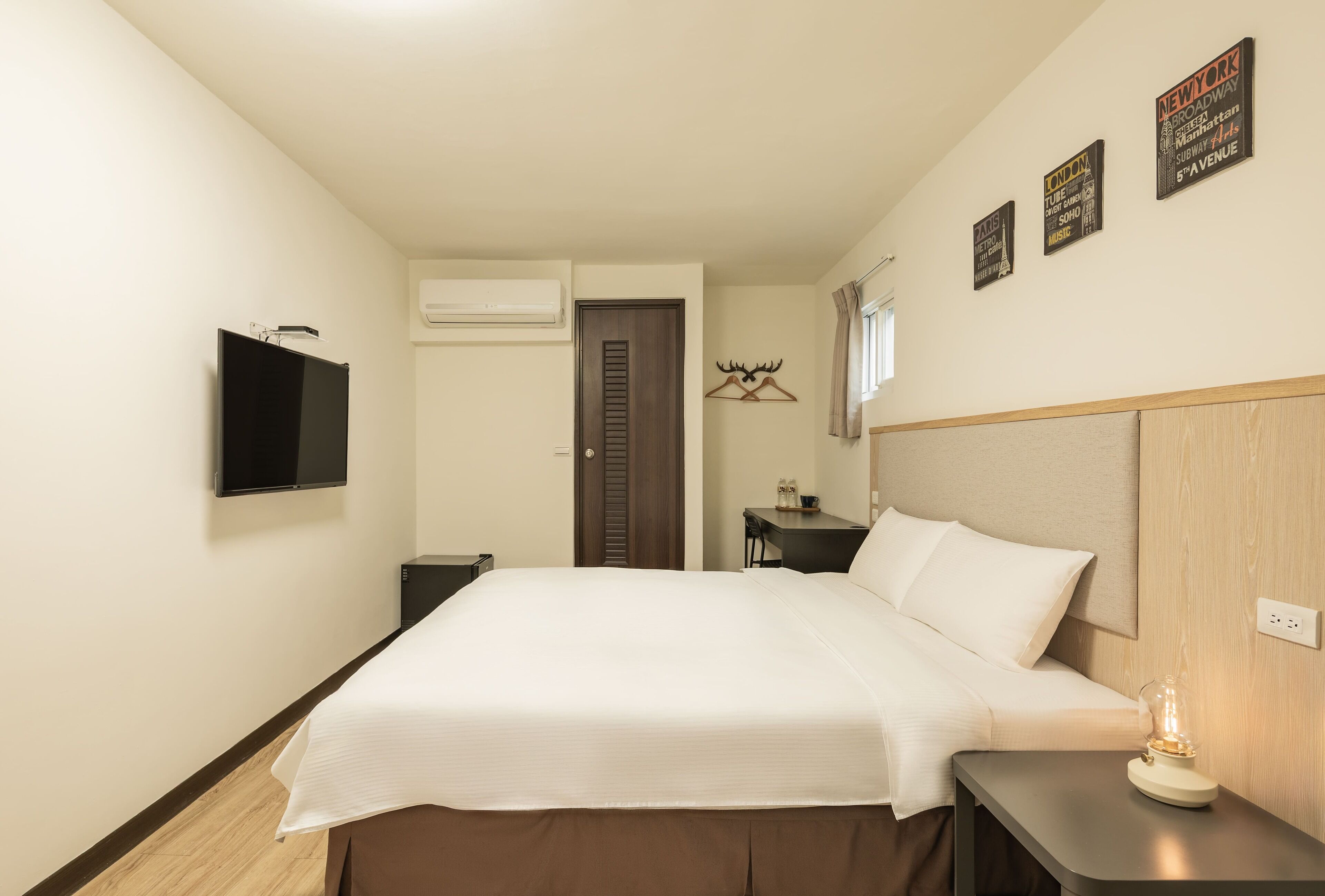 A hotel room with a bed, a bedside table with a lamp, a TV mounted on the wall, and a door leading to another room.