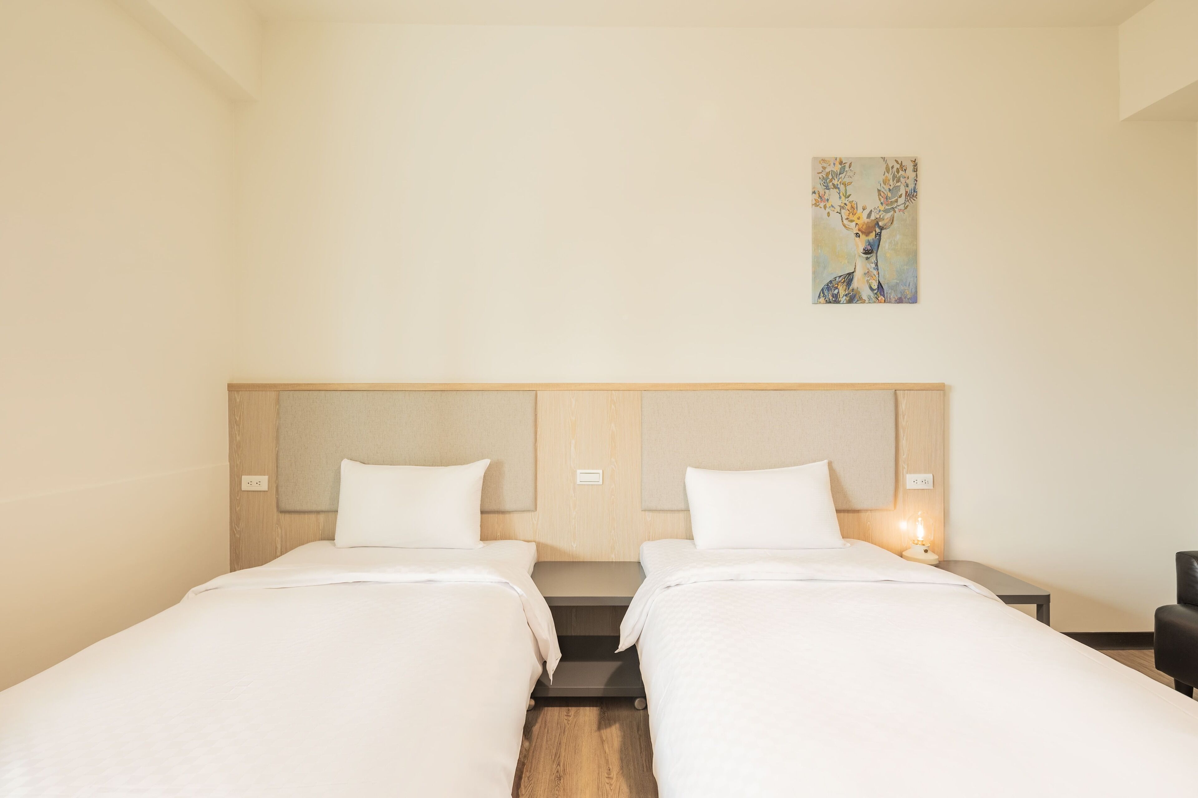 A hotel room with two single beds, a wooden headboard, a small painting on the wall, and a nightstand with a lamp.