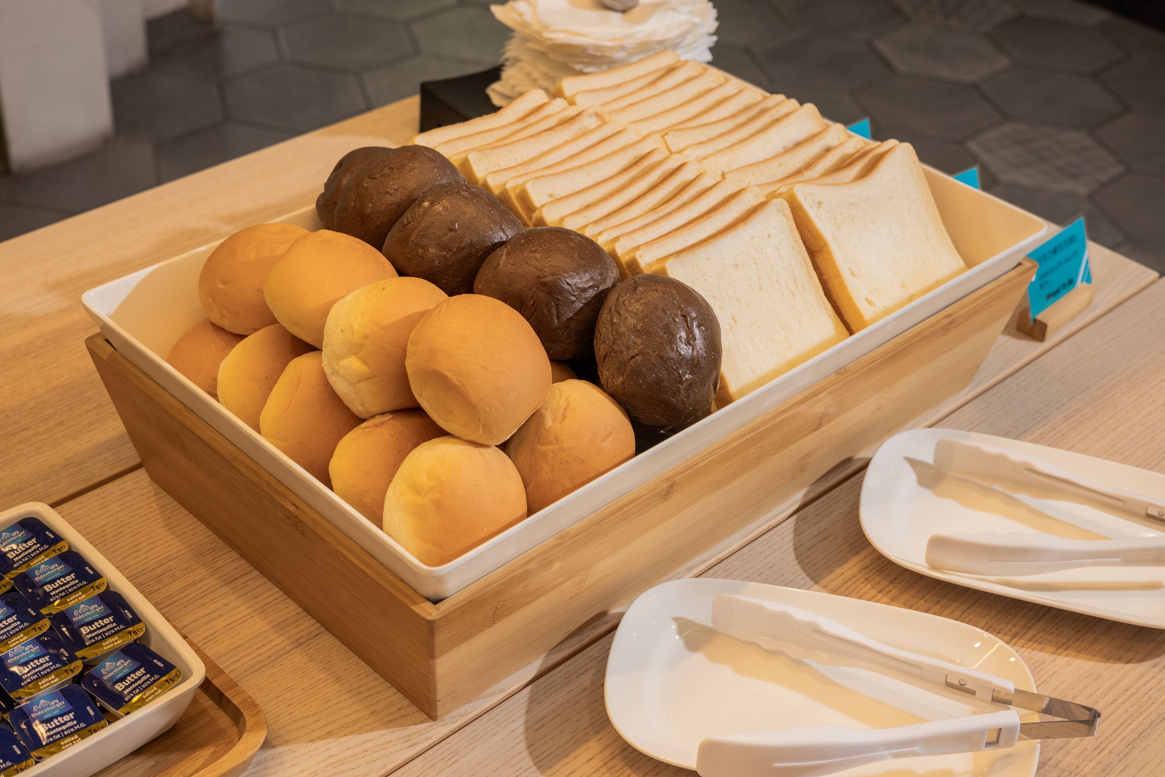 A wooden serving tray with various breads and rolls, a small container of butter, and white plates with utensils on a wooden table.