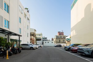 Un estacionamiento con varios coches y motocicletas, edificios con balcones y un cielo despejado.
