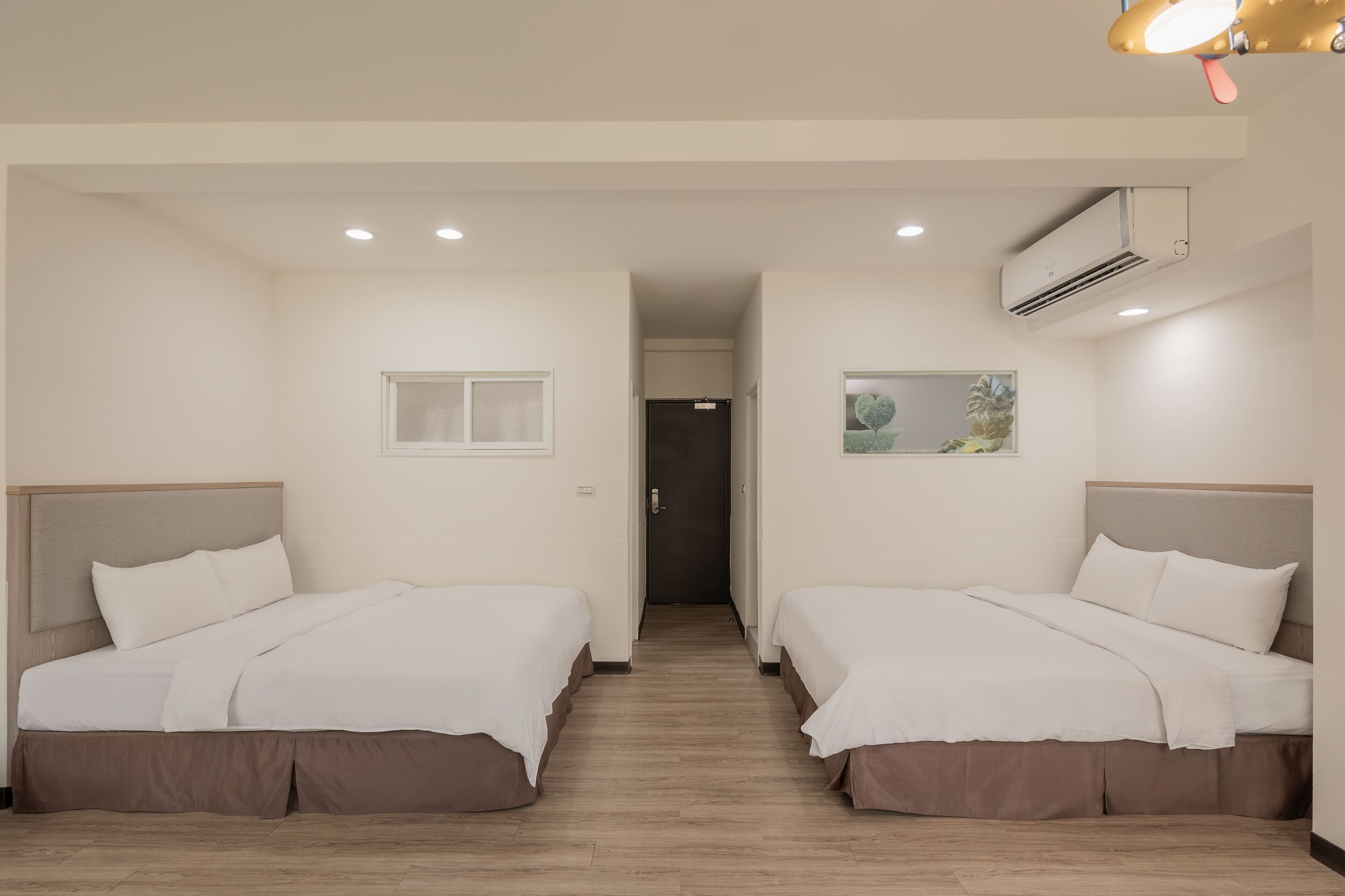 A hotel room with two beds, a window, an air conditioning unit, and a hallway leading to a door.