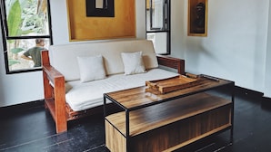 A modern living room with a sofa, a wooden coffee table, and a large window offering a view of greenery outside. - Montra Samui (Koh Samui)